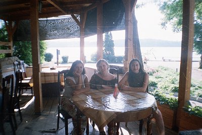 Three young women seated around a table covered with a floral tablecloth, framed by a wooden pergola. A scenic lakeside view ...