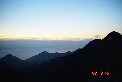 Dramatic, high-altitude landscape at twilight. Silhouetted mountain ridges descend towards a hazy, reflective body of water. ...