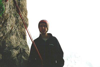 Rock climber, likely mid-climb, photographed with a blurred alpine background. He wears a red helmet, dark jacket & a beanie....
