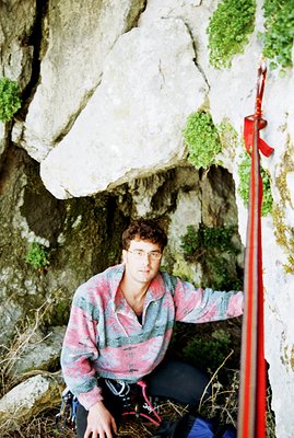 A climber, wearing glasses and a patterned fleece, poses within a rocky overhang. Equipped with climbing gear including ropes...