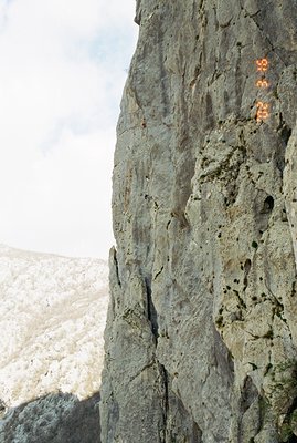 A lone rock climber ascends a sheer, grey cliff face, likely in an alpine setting. Snow-covered peaks are visible in the back...