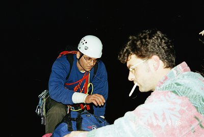 Two men, one wearing a climbing helmet, harness, and backpack, appear to be resting during a climbing activity. One man is sm...