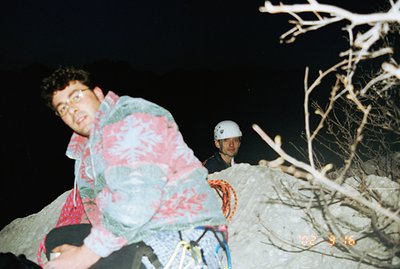 A climber, wearing a patterned puffer jacket and glasses, pauses on a rocky cliff face. Another climber is visible behind, we...