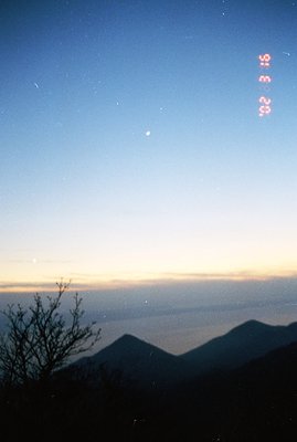 Dramatic twilight landscape with silhouetted mountains, visible sea, and a vibrant orange/yellow horizon. Sparse tree branche...