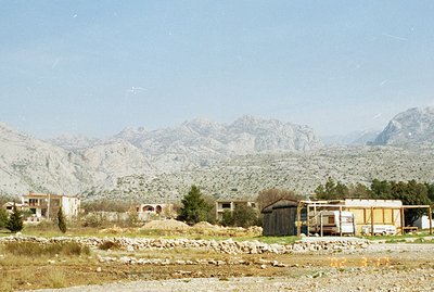 A view of rural buildings and a roadside structure set against a backdrop of dramatic, rocky mountains. Appears to be a trans...