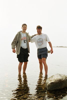 Two young men stand chest-deep in shallow, clear water, arms around each other, with a large rock visible at the water's edge...