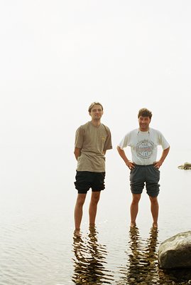 Two young men stand in shallow water, chest-deep, facing the camera. They wear casual summer attire: t-shirts and shorts. Gen...