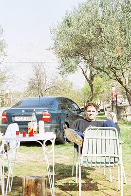 Man seated outdoors, appearing relaxed in a metal chair, near a dark green 1990s Mercedes sedan. The scene features a rustic,...