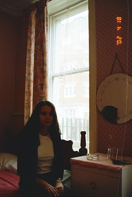 A young woman sits by a window with floral curtains, framed by textured wallpaper. View reveals a cityscape beyond. A circula...
