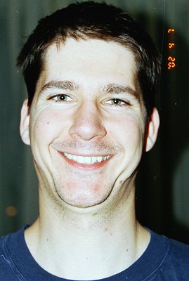 Close-up portrait of a young man smiling broadly. Short, dark hair and fair skin. Appears to be wearing a blue crew neck shir...