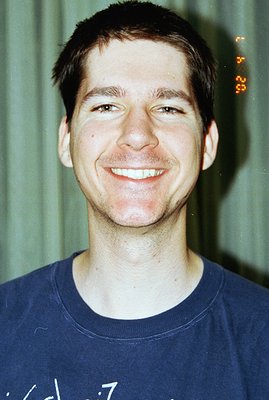 Close-up portrait of a young man with short dark hair, smiling broadly. He wears a blue t-shirt. The background appears to be...