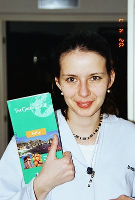 A woman holds a “Green Guide: Sicily” travel book and gives a thumbs-up. She's wearing a white zippered jacket and a dark bea...