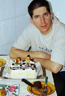 A young man stands proudly beside a decorated birthday cake with a playful lion figurine and inscription "Boldog születésnapo...