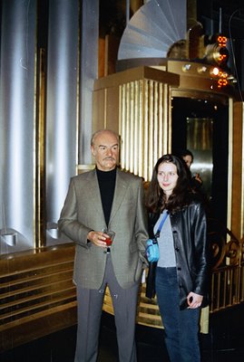 A distinguished gentleman in a tailored suit and turtleneck holds a drink in a grand, gold-accented lobby. A young woman in a...