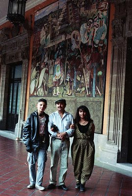 Three figures pose before a vibrant mural in what appears to be a grand, colonial-era courtyard. The mural depicts figures in...