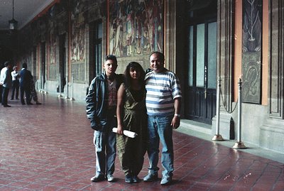 A family portrait showcases a young man, woman, and older man posed before a detailed mural depicting historical or religious...