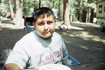 Close-up portrait of a young boy wearing a Los Angeles Lakers t-shirt, set against a backdrop of pine trees and an RV. Appear...