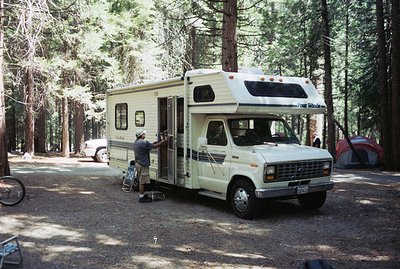 A vintage "Four Winds" Ford Econoline conversion van, likely from the 1980s, is parked in a forested campground. A man, weari...