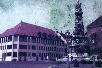 A vibrant, early-color photograph depicts a classical fountain monument & adjacent building with a red-tiled roof. The buildi...