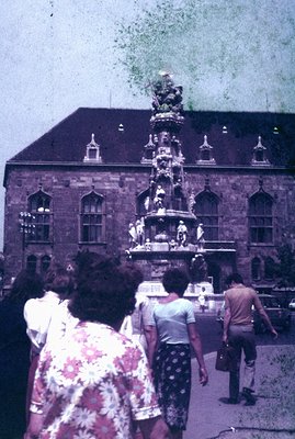 Detailed view of an ornate fountain set before a stately, multi-windowed building. Figures stroll by in casual summer attire;...