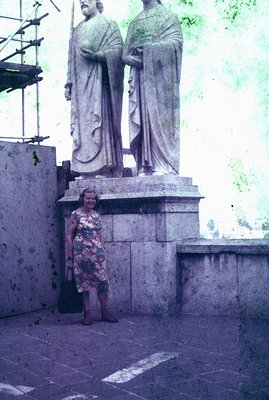 A woman stands near a large, classical-style stone statue of two figures, likely commemorating historical figures. She wears ...