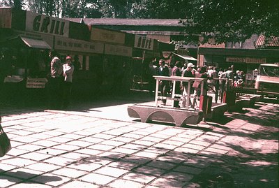 A low-angle view captures a vintage miniature train moving across a tiled plaza. Buildings with signage "Grill" and "Buffet" ...