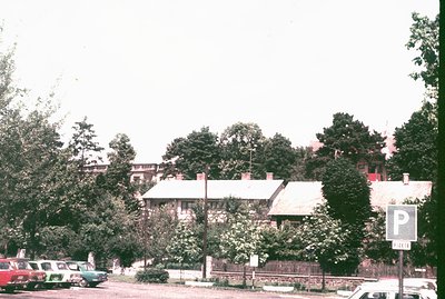 A roadside view showcases traditional, low-rise buildings with tiled roofs, likely residential. Lush trees obscure much of th...