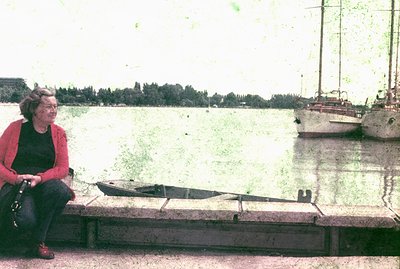 A woman sits on a stone embankment overlooking a body of water, likely a lake or bay. She wears a red cardigan and black pant...