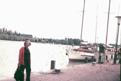 A woman stands by a dock next to a classic sailboat with tall masts. The scene features water, a distant shoreline with trees...