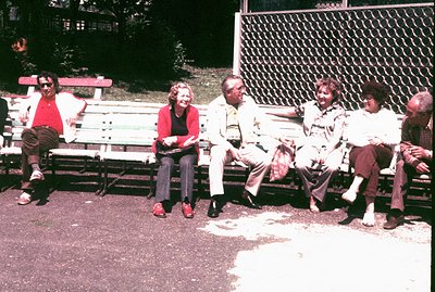 A group of five individuals are seated on a public park bench in what appears to be a casual, relaxed setting. The style of c...