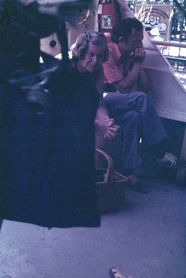 A young boy, circa 1970s, sits in jeans and a red t-shirt on a concrete ledge. A woman with long hair wears a dark dress & sa...