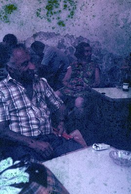 A candid shot captures two people seated at a table with a patterned tablecloth, likely in a café. The man, with a full beard...