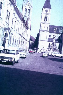 A wide, cobblestone street leads toward a towering church with a pointed steeple and ornate facade. Vintage cars, including a...