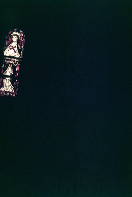 Fragment of a stained-glass window featuring a robed figure, likely a saint, depicted in profile. Vibrant blues, reds, and gr...