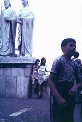 A candid street scene showing a young boy in casual clothing and two girls standing near a monumental statue, likely depictin...