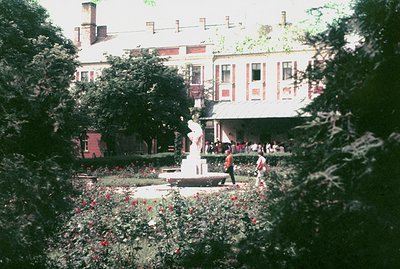 A formal garden with a central fountain featuring a classical statue, framed by dense foliage. A large, stately building with...
