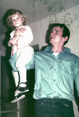 Man holds a young child aloft against a textured wall. Both wear casual clothing; the man in a light blue collared shirt and ...