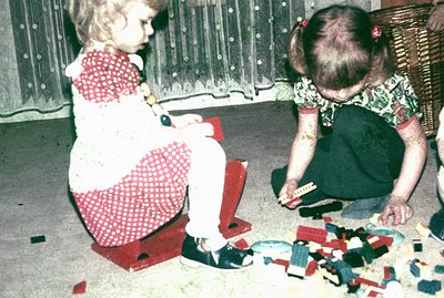 Two young girls play with toys in a warmly lit, domestic setting. One sits atop a red plastic toy, wearing a red-and-white pa...