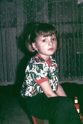 A young girl with a bowl cut sits indoors, gazing directly at the camera. She wears a vibrant, patterned shirt with red trim ...