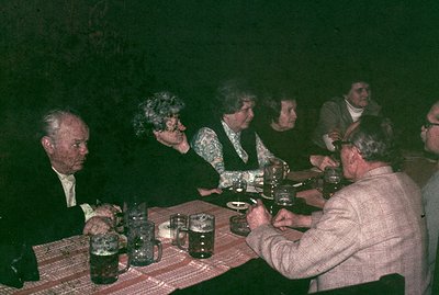 A group gathers around a table, likely enjoying a meal or drinks. Visible are several older adults; men in suits and sweaters...