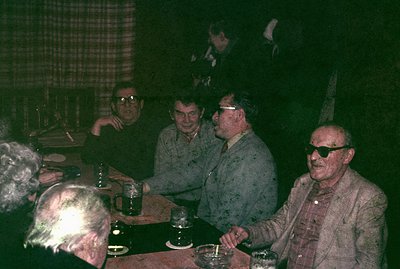 Four men sit around a table in a dimly lit, likely indoor setting. Several glasses and ashtrays are visible. The group’s atti...