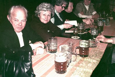 A group gathered around a table with steins of beer, possibly in a German beer garden or similar establishment. A couple, lik...