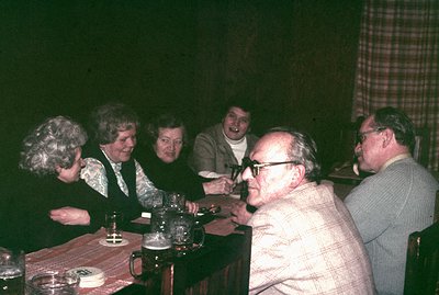 A group of five adults gathered around a wooden table in a dimly lit room. Floral and patterned attire suggest a casual, home...