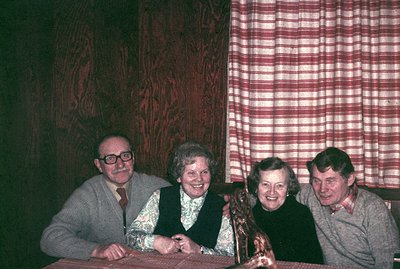 Four individuals seated at a table covered with red fabric, posed for a portrait. The décor includes wood-paneled walls and a...