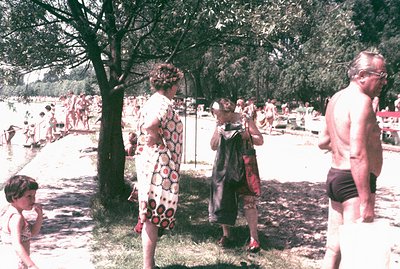 A lakeside scene with families enjoying a summer day. A young girl stands near a woman in a 1960s-70s patterned dress & hat. ...