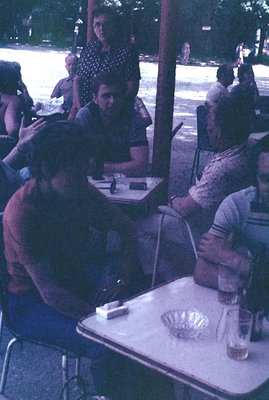 Three men seated at a table, appearing to converse in an outdoor setting. One man has dark hair, the others are lighter-haire...