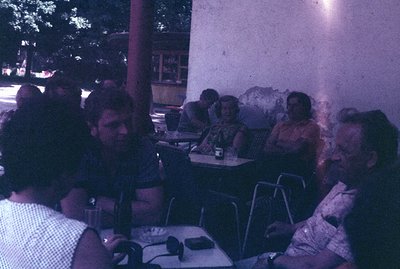 Group of men and women seated at tables outdoors; likely a cafe or restaurant patio. The scene shows casual clothing, hairsty...