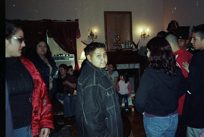 A group gathers in what appears to be a grand, older home. A young boy in a denim jacket stands facing the camera. The room f...