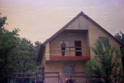 A two-story building, seemingly residential, features a prominent balcony with a single figure visible. The architecture sugg...