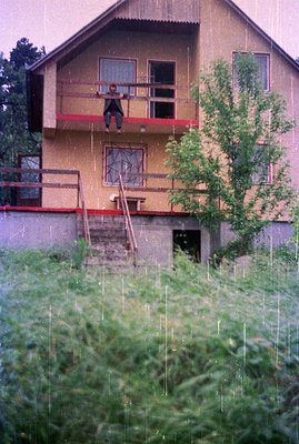 A two-story building with a balcony featuring a person seated and facing away. Visible architectural details include a red-tr...